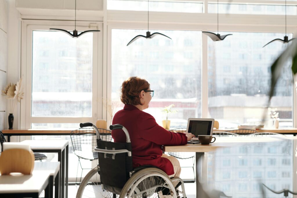 A woman in a red blazer works on a laptop in a bright café, seated in a wheelchair.