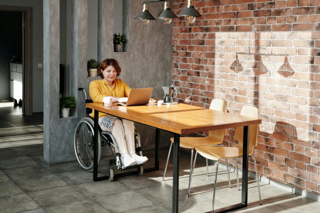 A woman in a wheelchair works on her laptop from home in a modern, sunny interior.