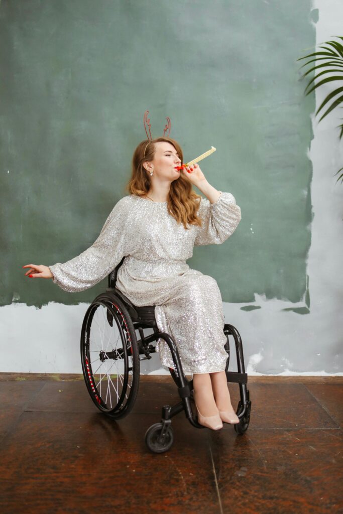 Joyful woman in a wheelchair wearing reindeer headband and blowing party horn indoors.